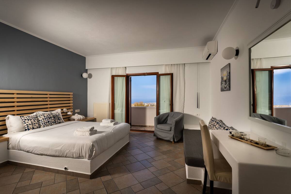 A modern hotel room with a large bed, a desk, a chair, and a balcony with a view.