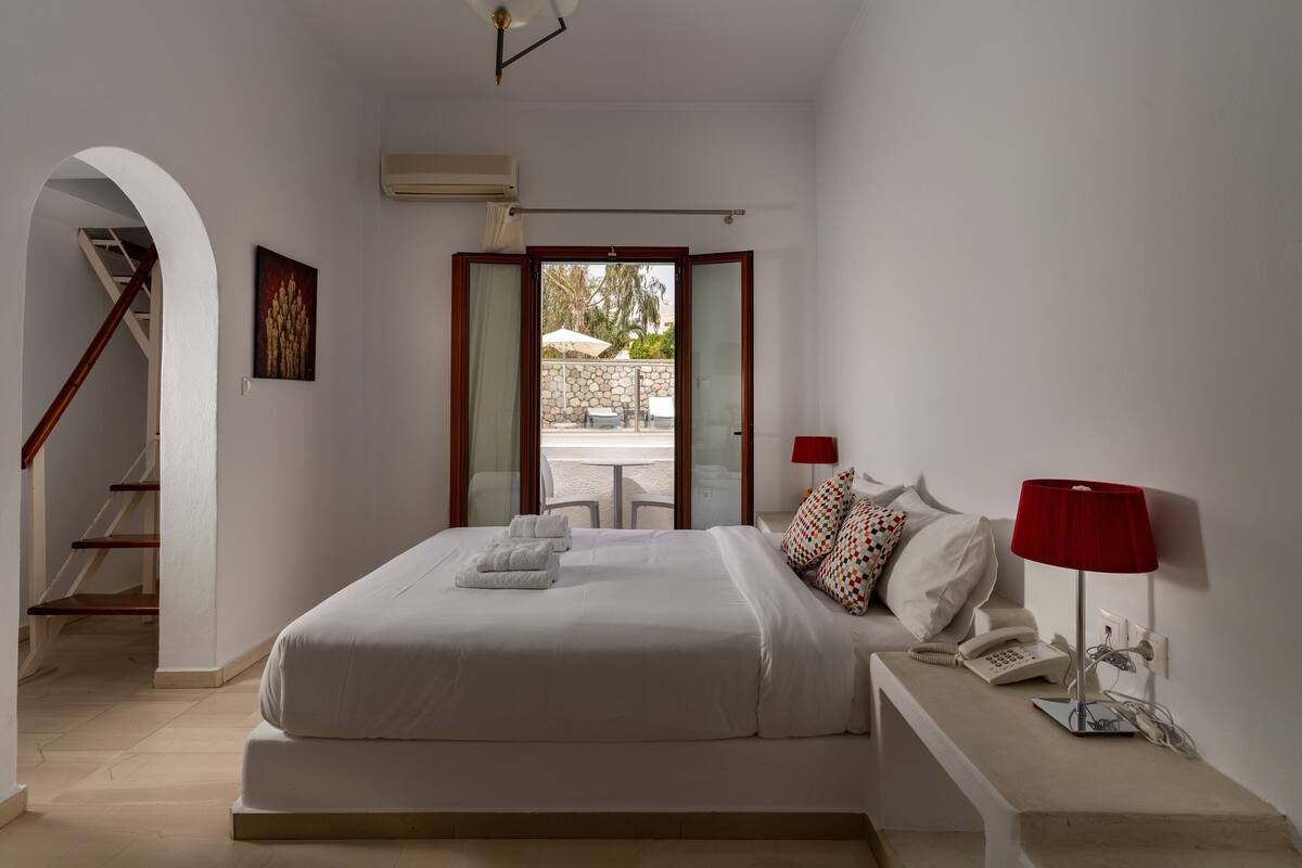 A hotel room with a bed, a desk with a lamp, a phone, and a view of the beach through a glass door.