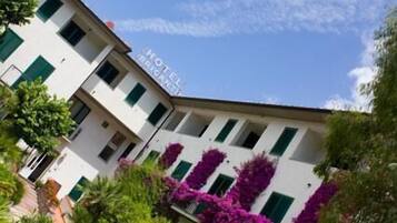 Un hotel con facciata bianca, persiane verdi e piante fiorite di colore viola davanti.