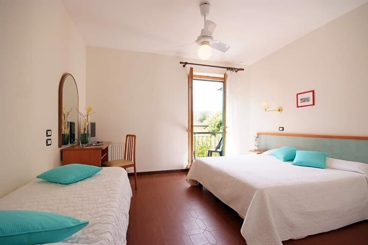 Une chambre d’hôtel avec deux lits, un ventilateur de plafond, une fenêtre avec vue, un bureau avec une chaise et une petite table avec une lampe.