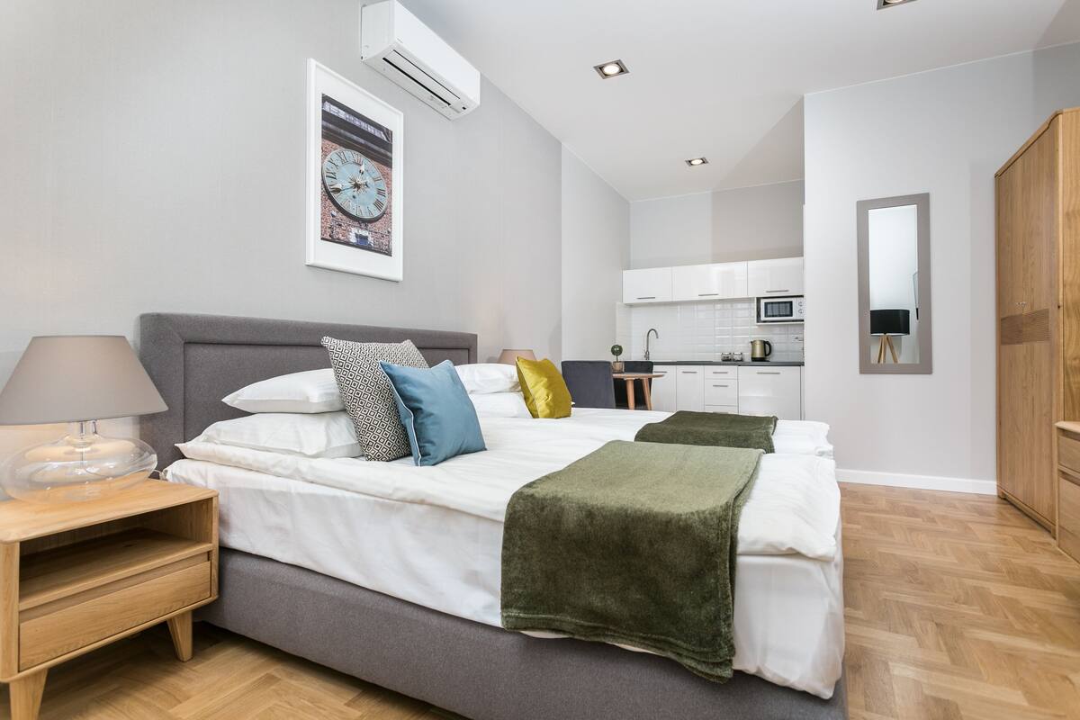 Un dormitorio moderno con una cama grande, mesitas de noche, un tocador y una zona de cocina al fondo.