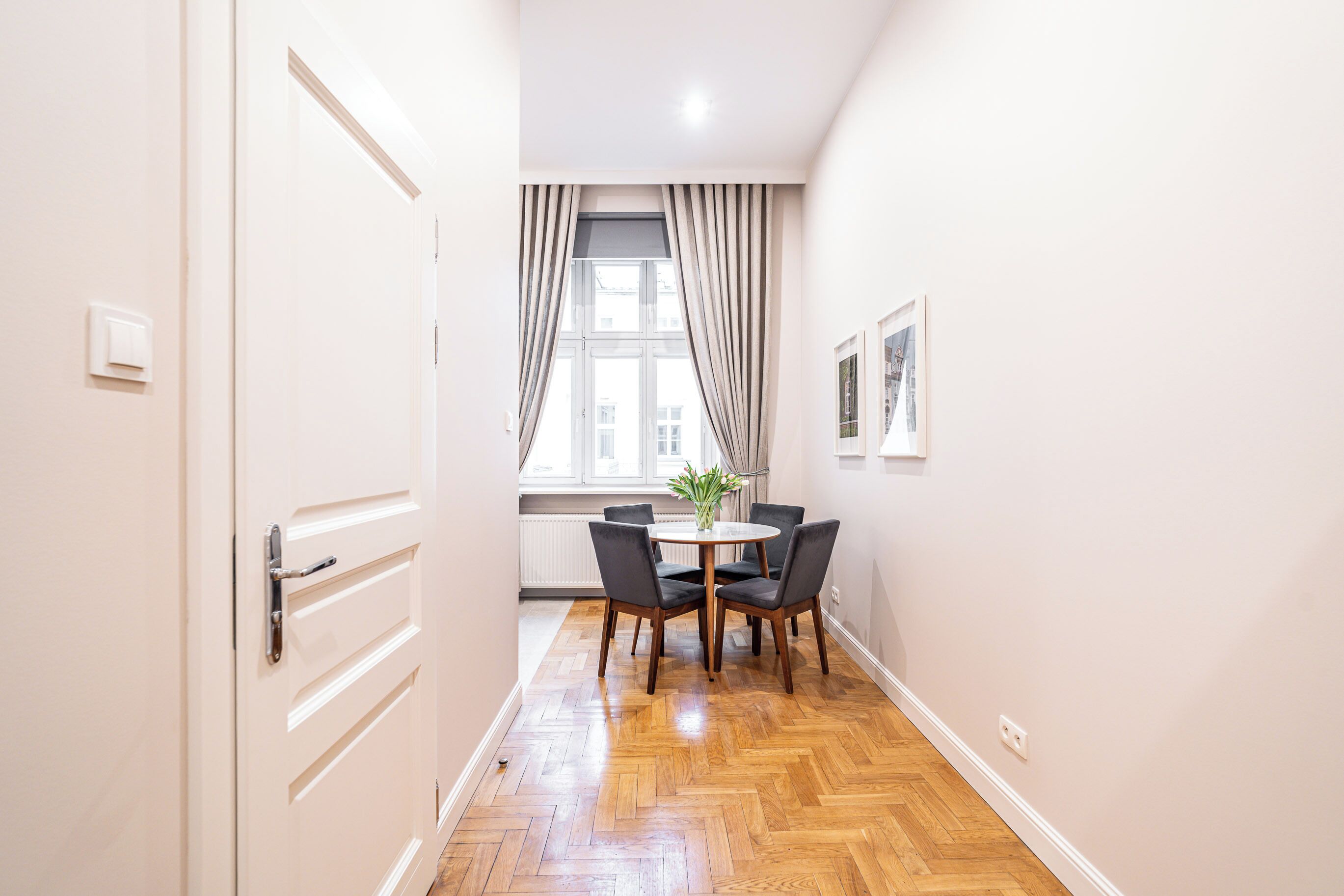 A hallway with a wooden floor, a white door, a window with curtains, and a table with chairs.