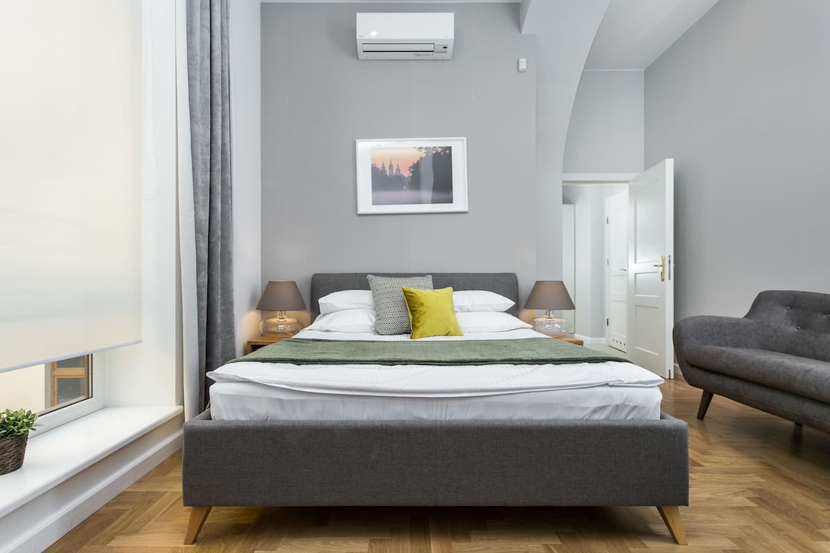 Un dormitorio moderno con una cama grande, lámparas de noche, un sofá gris, una ventana con vistas y un cuadro en la pared.