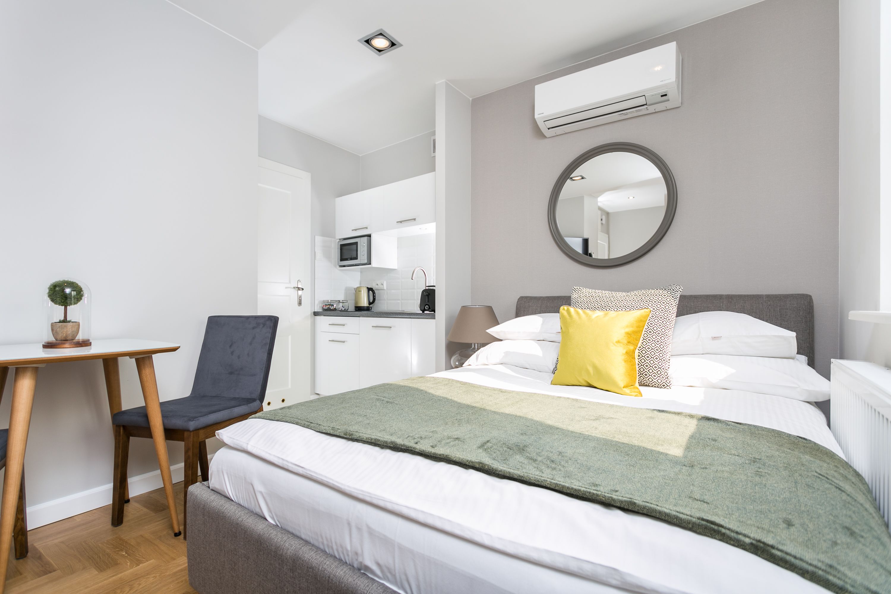 A modern hotel room with a bed, a desk, a chair, a round mirror, and a kitchenette in the background.