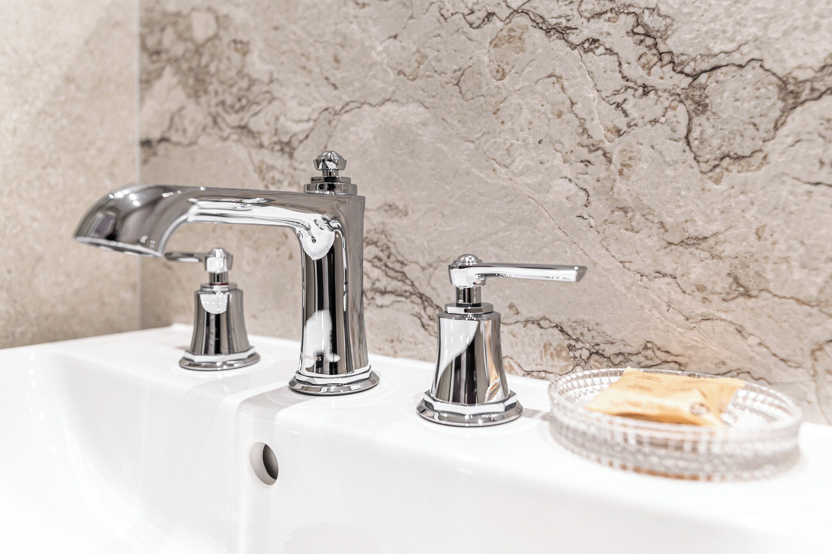 A bathroom with a white sink, two chrome faucets, and a marble wall.