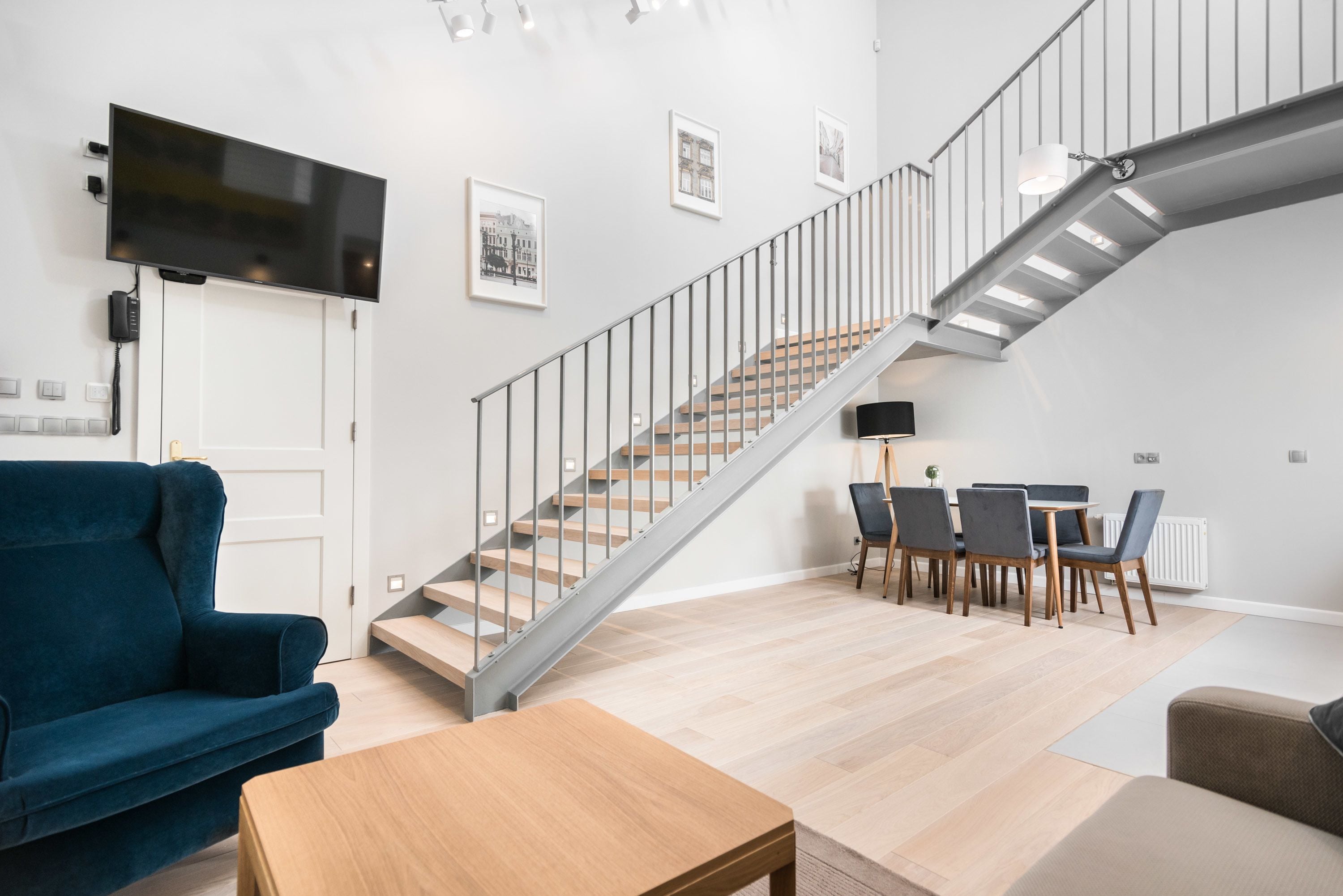 A modern living room with a staircase, a flat-screen TV mounted on the wall, and a dining area with chairs and a table.