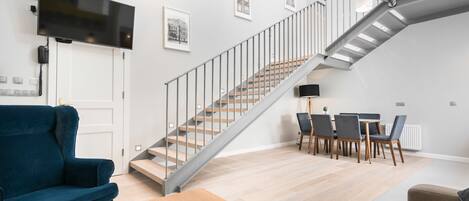 A modern living room with a staircase, a flat-screen TV mounted on the wall, and a dining area with chairs and a table.