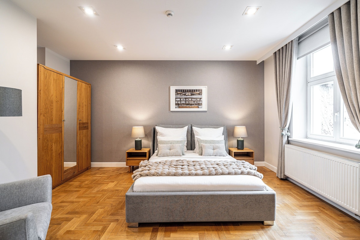 Quarto com uma cama grande, guarda-roupa de madeira, duas mesas de cabeceira com luminárias, um quadro emoldurado na parede e uma janela com cortinas.