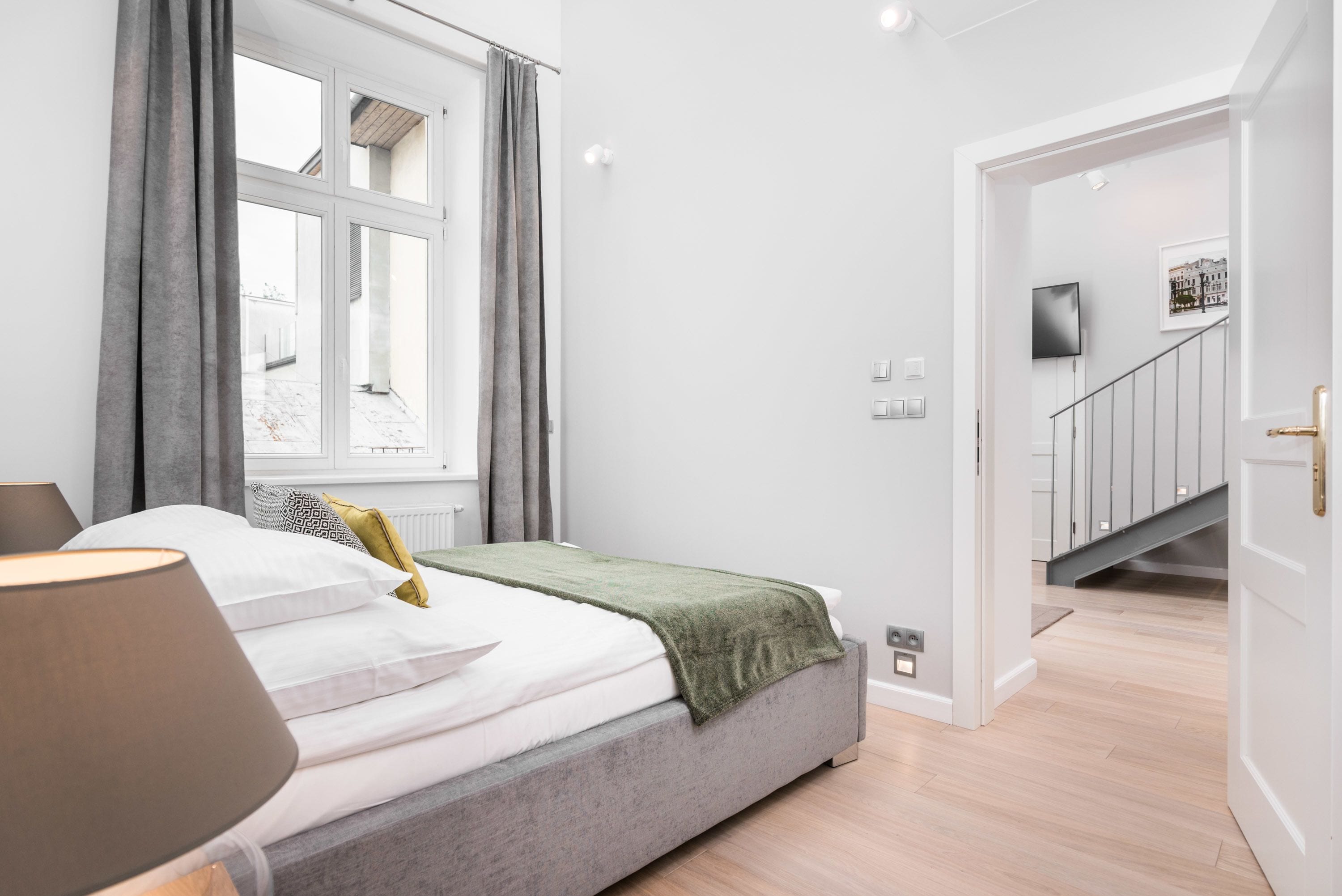 Une chambre à coucher avec un lit, une fenêtre avec des rideaux, une lampe et un escalier menant à une autre pièce.