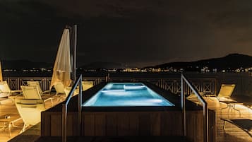 An outdoor swimming pool with lounge chairs and umbrellas, surrounded by a wooden deck and railing, overlooking a body of water and distant hills.