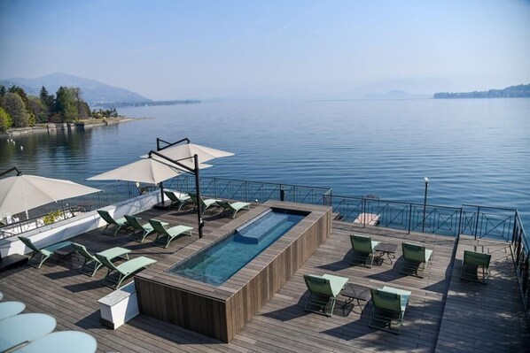 A deck with a pool, lounge chairs, and umbrellas overlooking a lake. - Hotel Bel Sit (Meina)