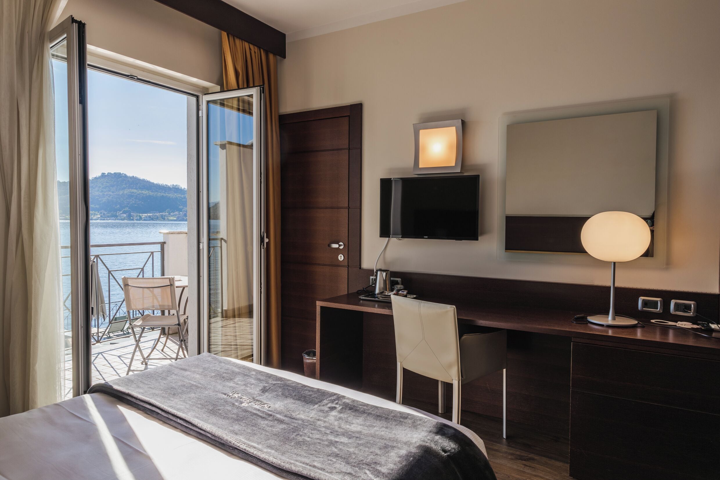 A hotel room with a bed, a desk, a chair, a TV, a lamp, a balcony with a view of the sea, and a window with curtains.