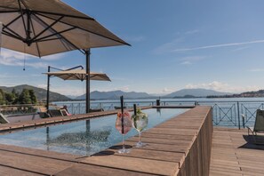 A wooden deck with a pool, two umbrellas, and a view of mountains and water. - Hotel Bel Sit (Meina)