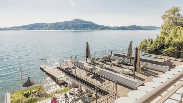 Une terrasse au bord du lac, dotée de chaises longues, d’une piscine et d’un espace repas offrant une vue sur l’eau.