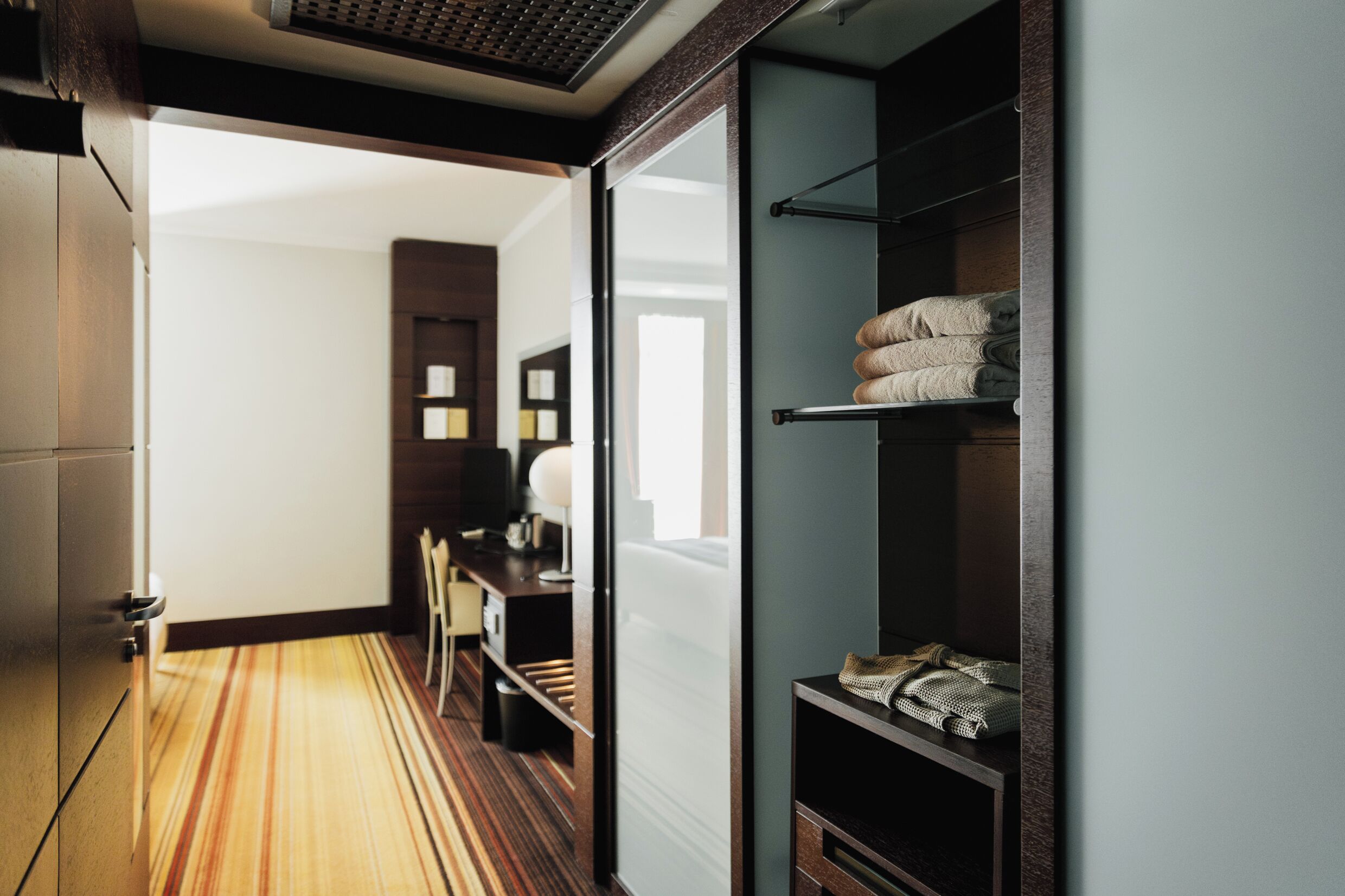 A hotel room with a wooden floor, a desk with a chair, a bed with a headboard, and a wardrobe with folded towels.