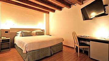 Quarto com cama, mesa de cabeceira, escrivaninha, cadeira e televisão fixada na parede.