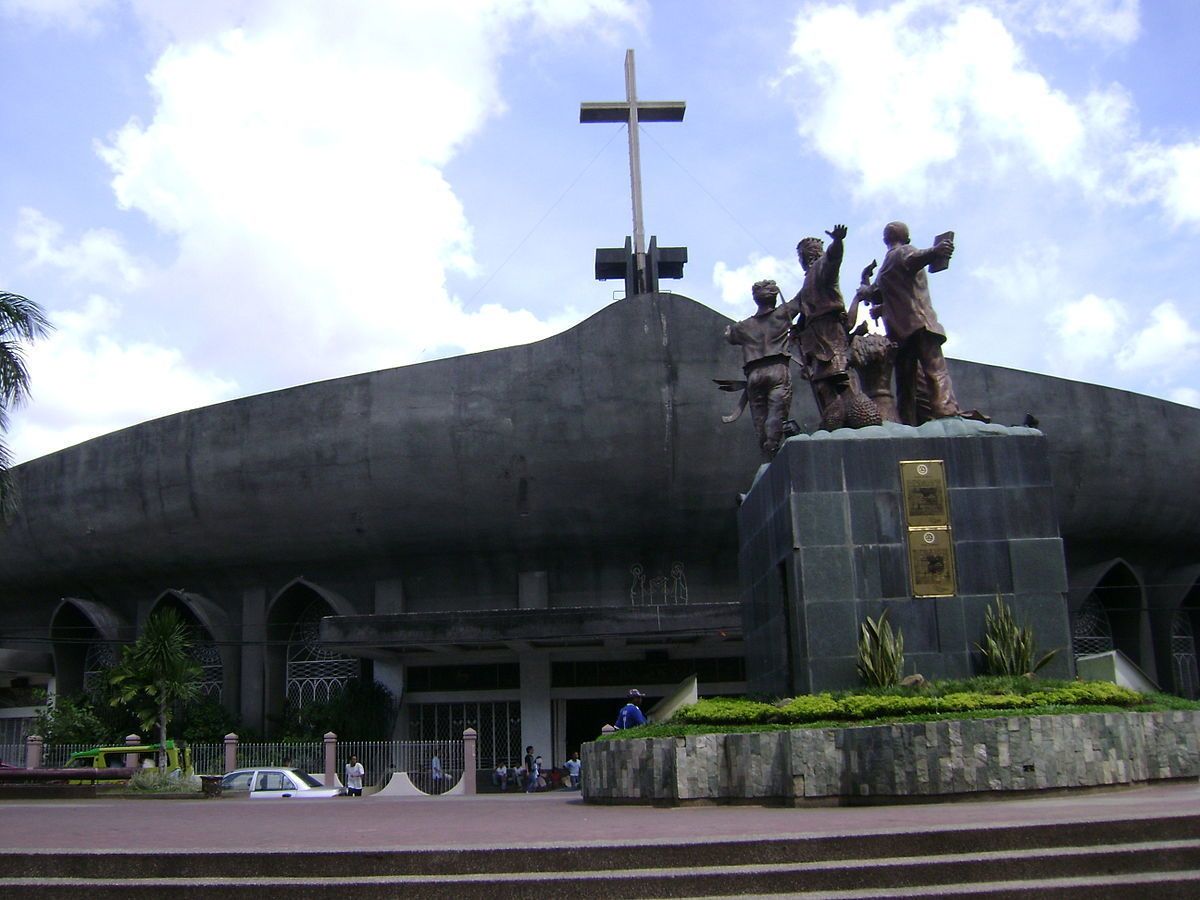 Um edifício grande com uma cruz no topo, uma estátua em frente e algumas pessoas por perto.