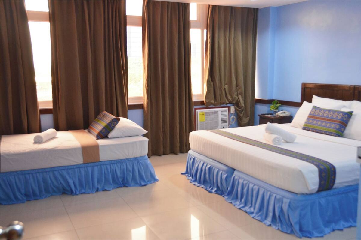 A hotel room with two beds, each with a blue skirted bedspread, a white pillow, and a rolled towel. There is a small table with a plant and a phone on it.