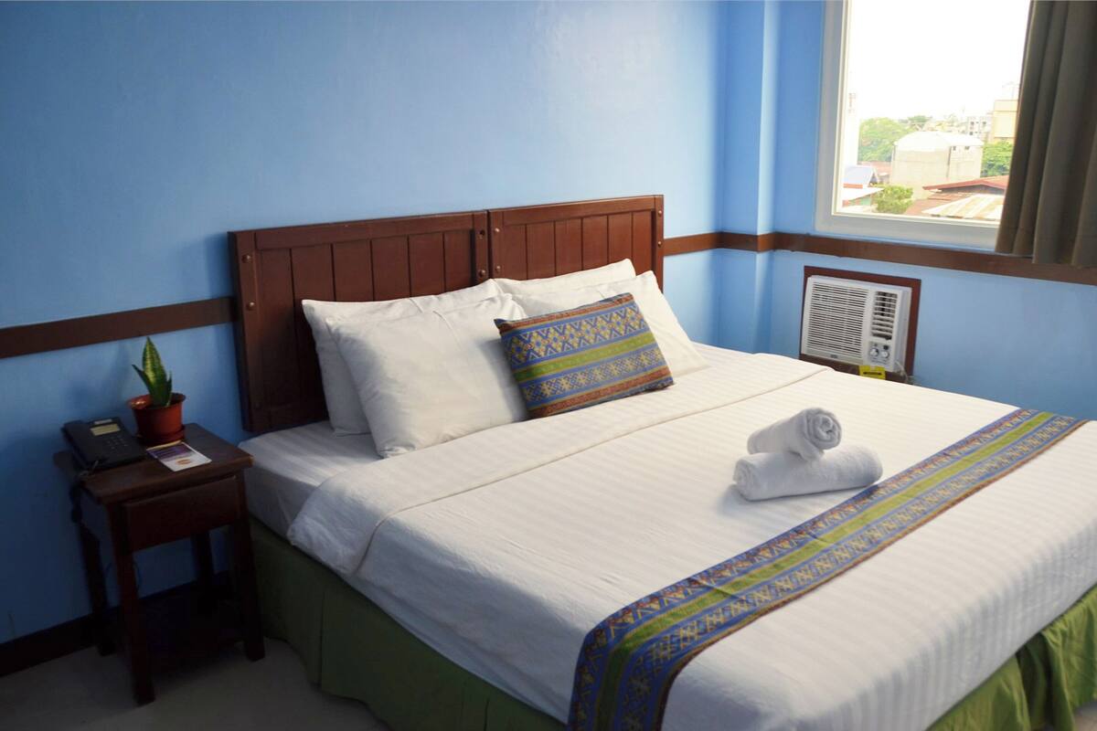 A neatly made bed with white linens, a patterned pillow, and a rolled towel on the bed. There is a small wooden nightstand with a telephone and a potted plant beside the bed.