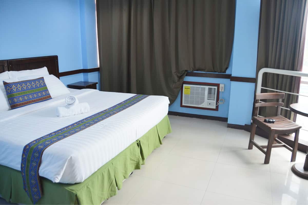 A hotel room with a neatly made bed, a green skirted bedspread, a wooden nightstand, a chair, and a window with curtains.