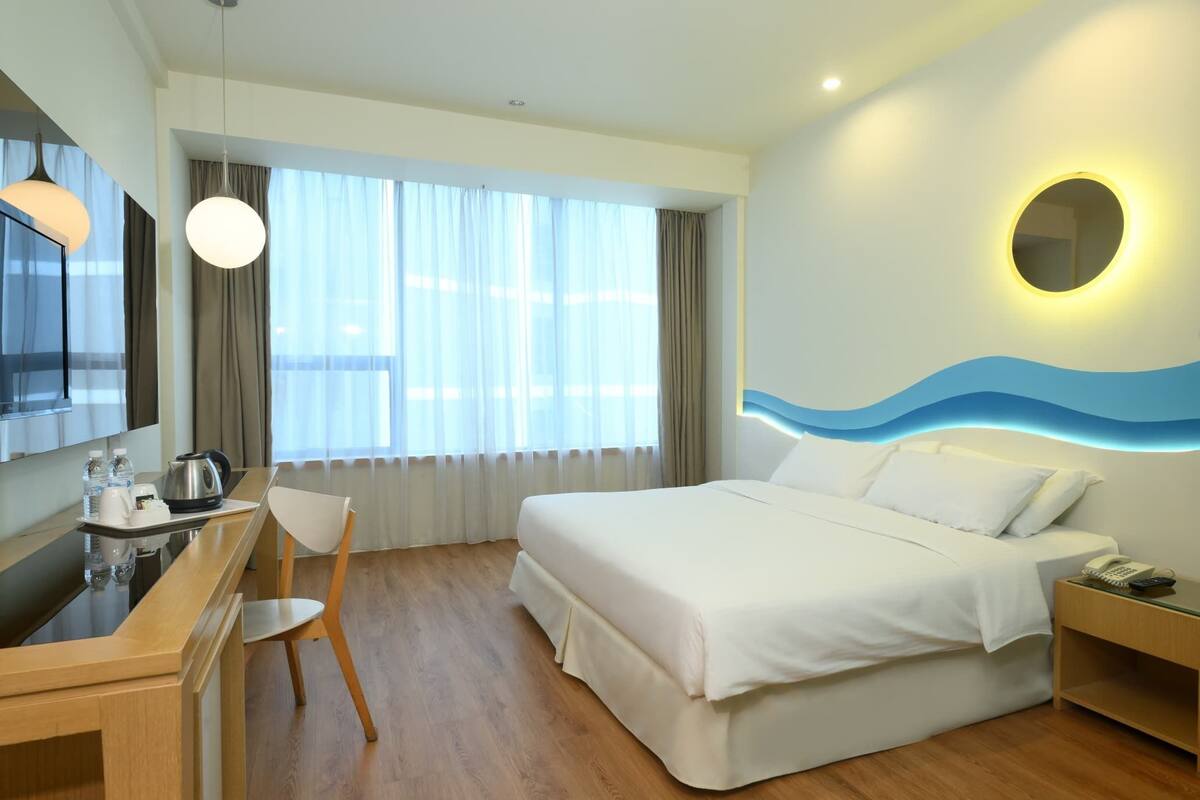 A hotel room with a bed, bedside table, chair, desk, and a large window with curtains.