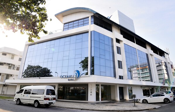 A modern hotel building with a glass facade, a white van parked in front, and a car parked to the side. - Oceania Hotel (Kota Kinabalu)
