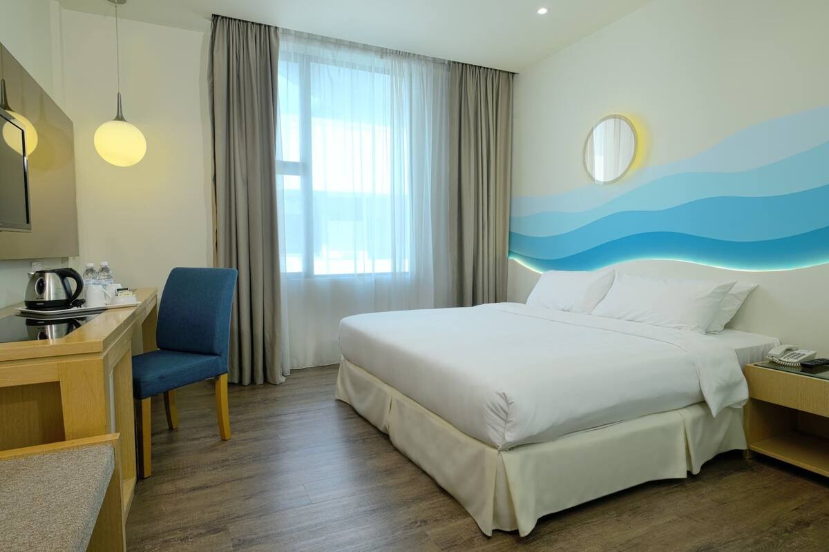 A hotel room with a bed, a desk with a chair, a mirror, and a view of the outdoors through a window.