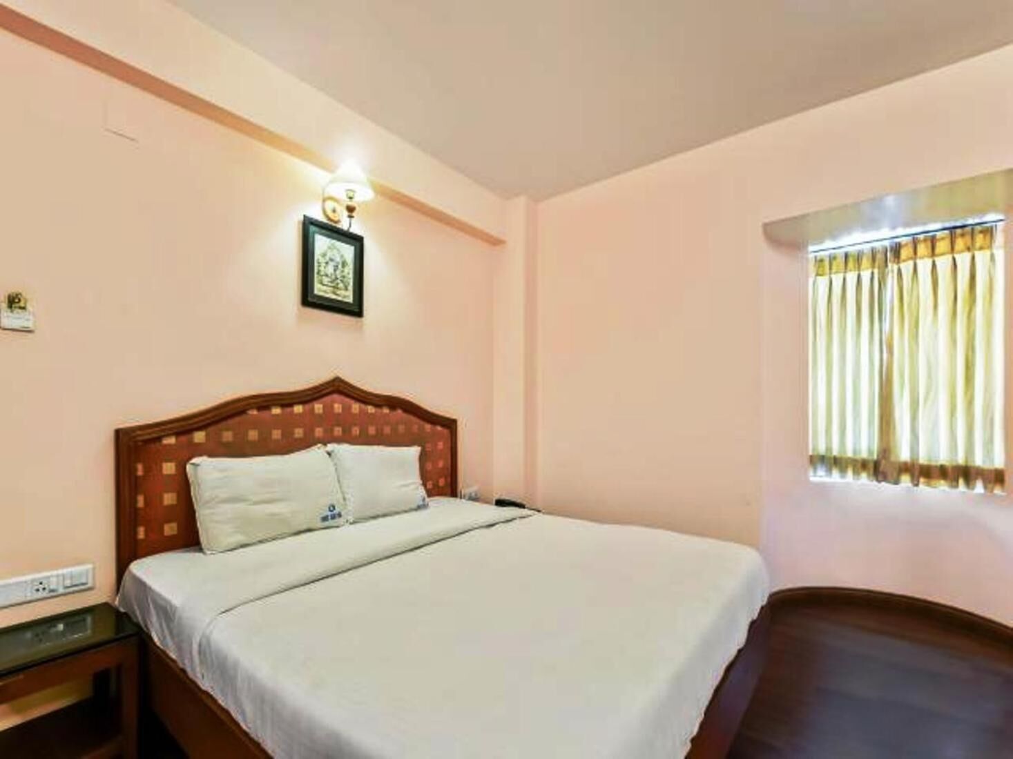 A hotel room with a single bed, a bedside table, a wall-mounted light, and a window with curtains.