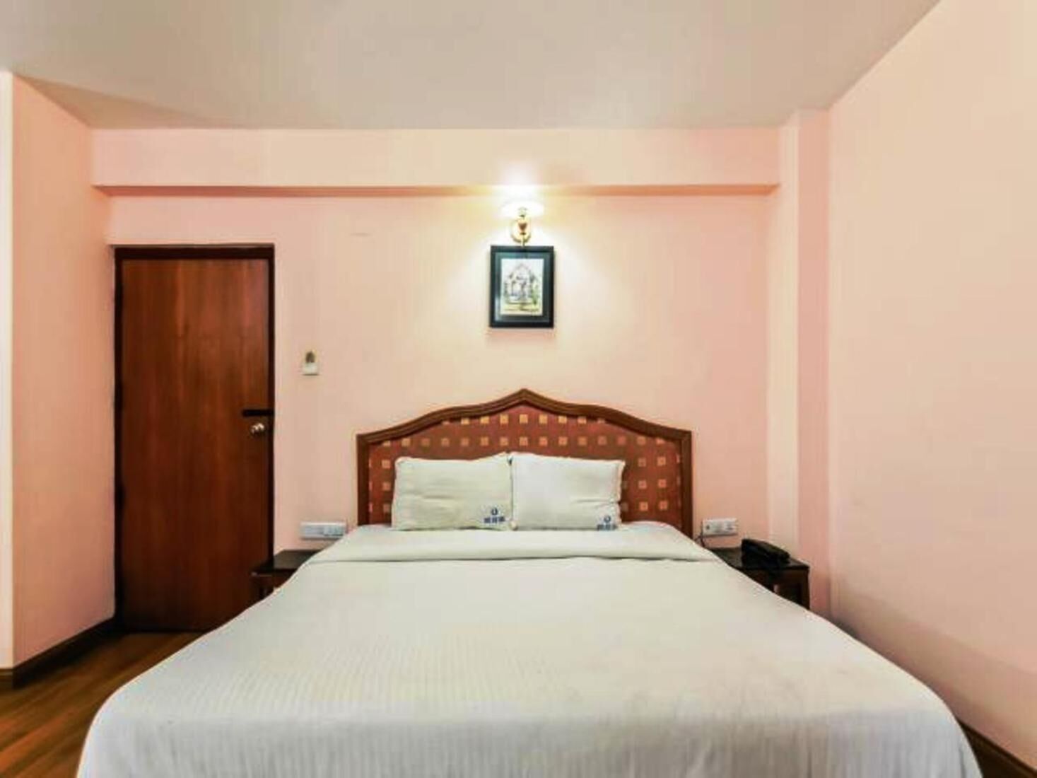 A bedroom with a wooden headboard, a bed with white bedding, a nightstand, a framed picture on the wall, and a door.