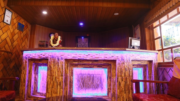 A room with a wooden ceiling, a stone wall with a purple light, a framed certificate, and a window with a view. - Hotel Blue Bird (Ootacamund)
