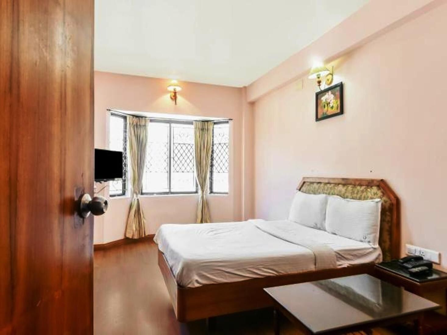 A hotel room with a wooden bed, a bedside table, a flat-screen TV, and a window with curtains.