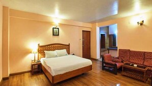 A bedroom with a wooden bed, a nightstand, a red patterned sofa, and a small table. - Hotel Blue Bird (Ootacamund)
