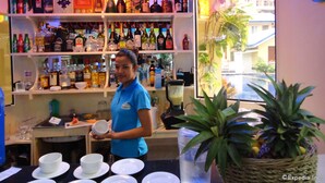 A bar with shelves of liquor bottles, a woman holding a cup, and a window with a view of a building and a pool. - Blue Lagoon Inn & Suites (Puerto Princesa)