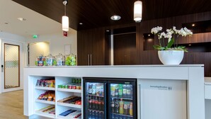 A modern reception area with a white counter, refrigerated beverage dispensers, and a potted orchid. - Hampton by Hilton Amsterdam / Arena Boulevard (Amsterdam)