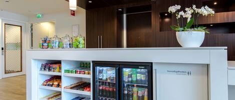 Una moderna zona de recepción con un mostrador blanco, dispensadores de bebidas refrigeradas y una orquídea en maceta.