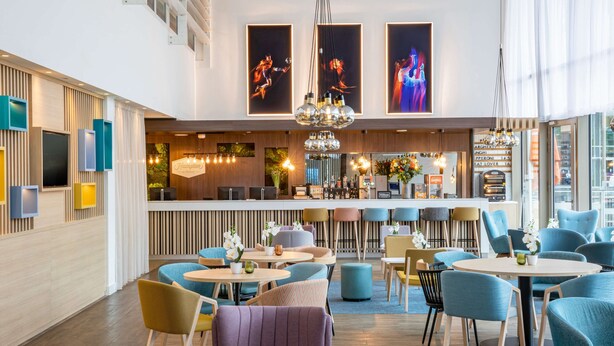 A modern hotel lobby with a bar area, seating arrangements, and artwork on the walls. - Hampton by Hilton Amsterdam / Arena Boulevard (Amsterdam)