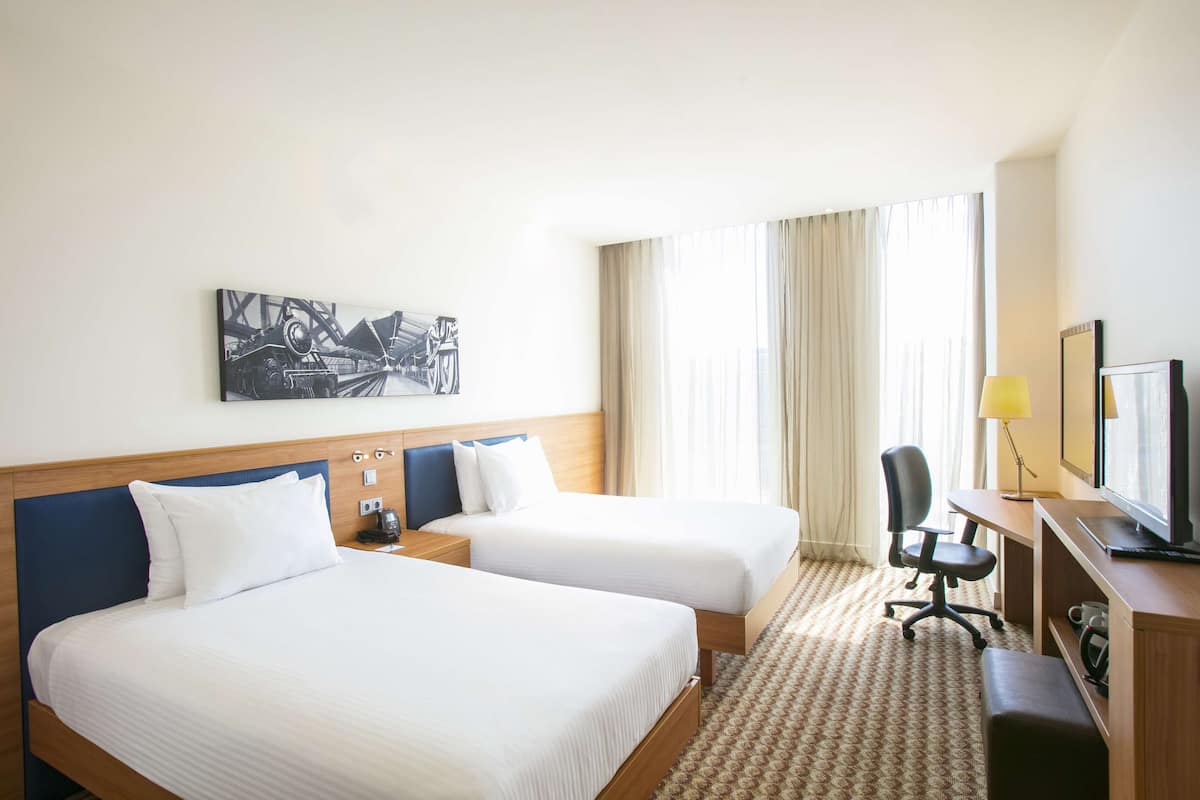 Ein Hotelzimmer mit zwei Betten, einem Schreibtisch, einem Stuhl, einem Fernseher und einem Bild an der Wand.