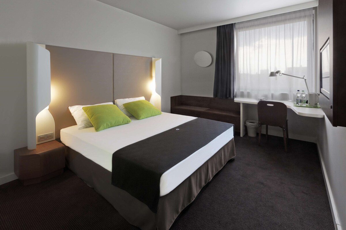 A hotel room with a bed, bedside table, desk, chair, and a window with curtains.