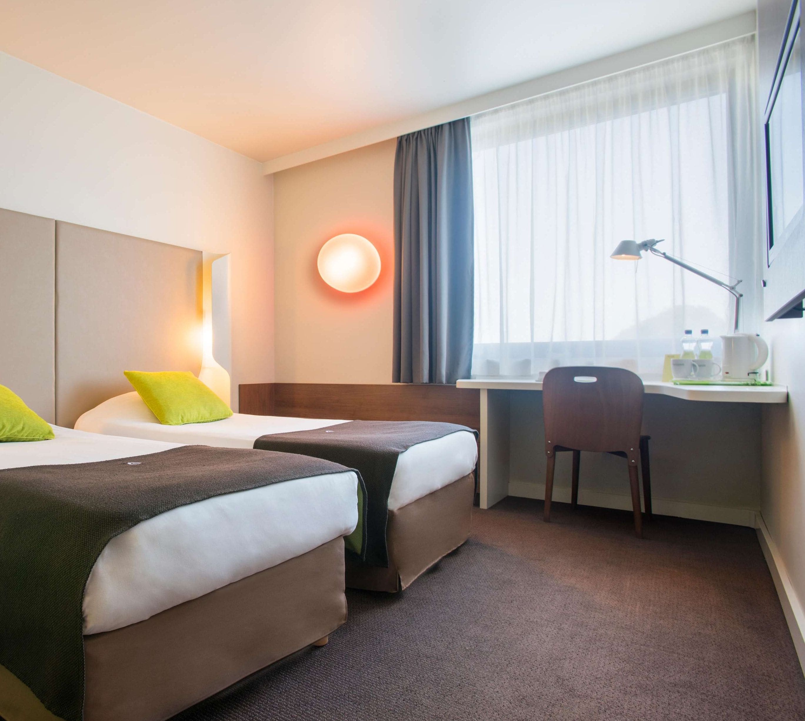 Habitación de hotel con dos camas, un escritorio, una silla, una lámpara y una ventana con cortinas.