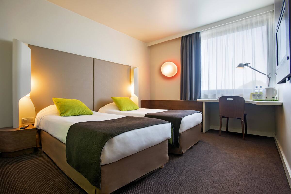 A hotel room with two beds, a desk, a chair, and a large window with curtains.