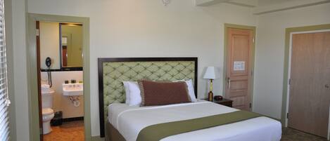 A hotel room with a large bed, a bedside table, a mirror, a bathroom with a toilet and sink, and a door.