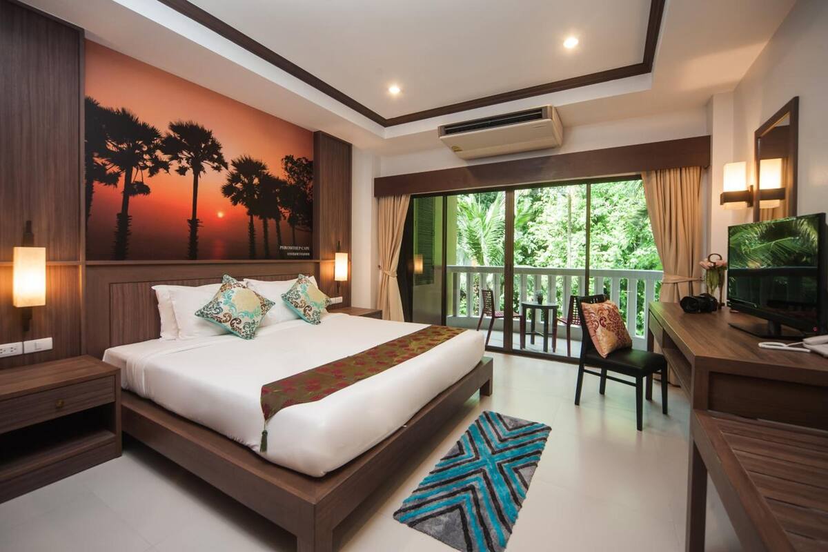 Quarto de hotel com uma cama grande, uma mesa de trabalho com televisão, varanda com vista para vegetação e um mural de pôr do sol na parede.