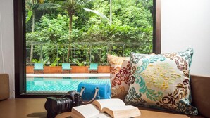 A view of a swimming pool with lounge chairs, a blue suitcase, a camera, and an open book on a table by a window with a patterned pillow.