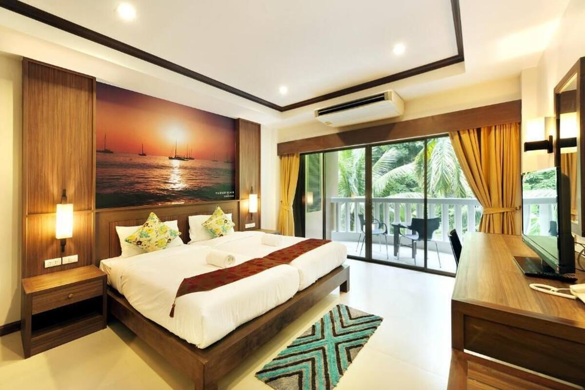 Um quarto espaçoso com uma cama grande, uma pintura de um pôr do sol sobre a água, uma varanda com vista para palmeiras e uma escrivaninha com telefone.