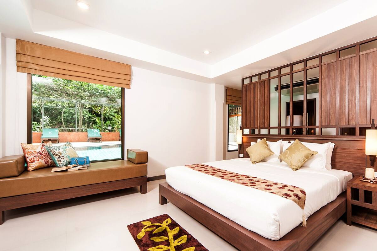 Quarto moderno com cama grande, um banco embutido, mesa de cabeceira e vista para uma piscina.