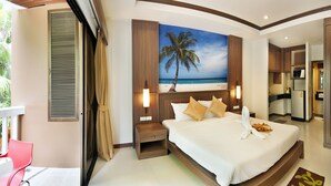 A hotel room with a large bed, a painting of a beach scene, a small kitchen area, and a balcony with a red chair.