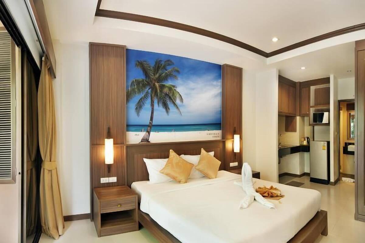 Quarto de hotel com cama, mesa de cabeceira, pintura de uma paisagem praiana, cozinha compacta e banheiro.