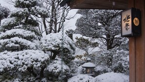 A snow-covered garden with traditional Japanese architecture, including a lantern and a wooden structure with Japanese characters.