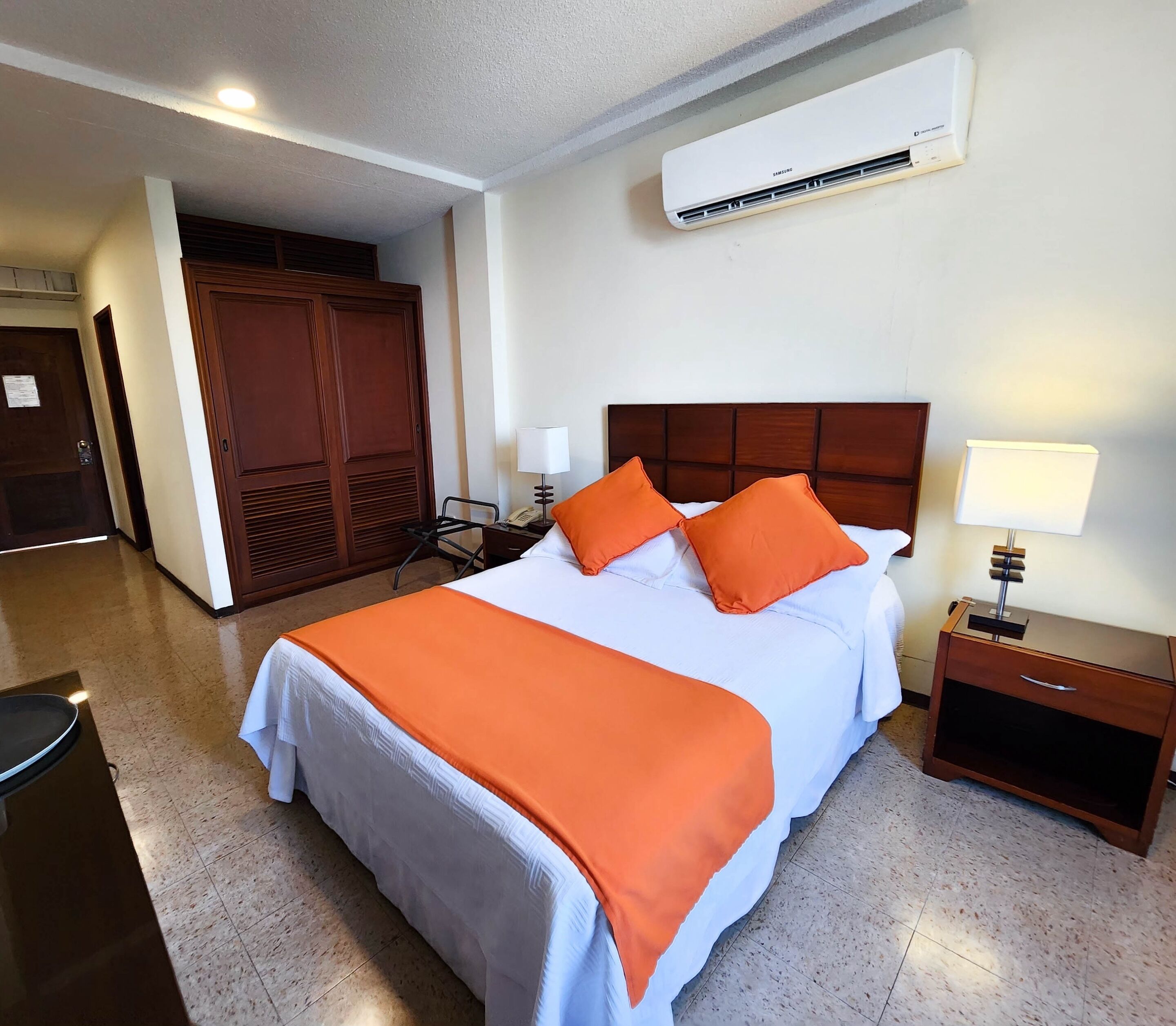 Ein Hotelzimmer mit Bett, Klimaanlage, Nachttisch, Lampe und Kleiderschrank.