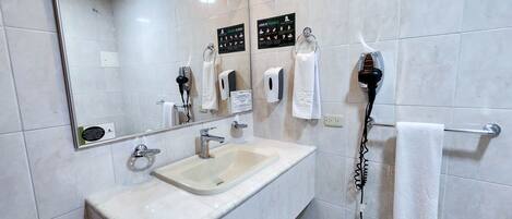 Un baño con lavamanos, espejo, toallero, secador de manos y dispensador de jabón.